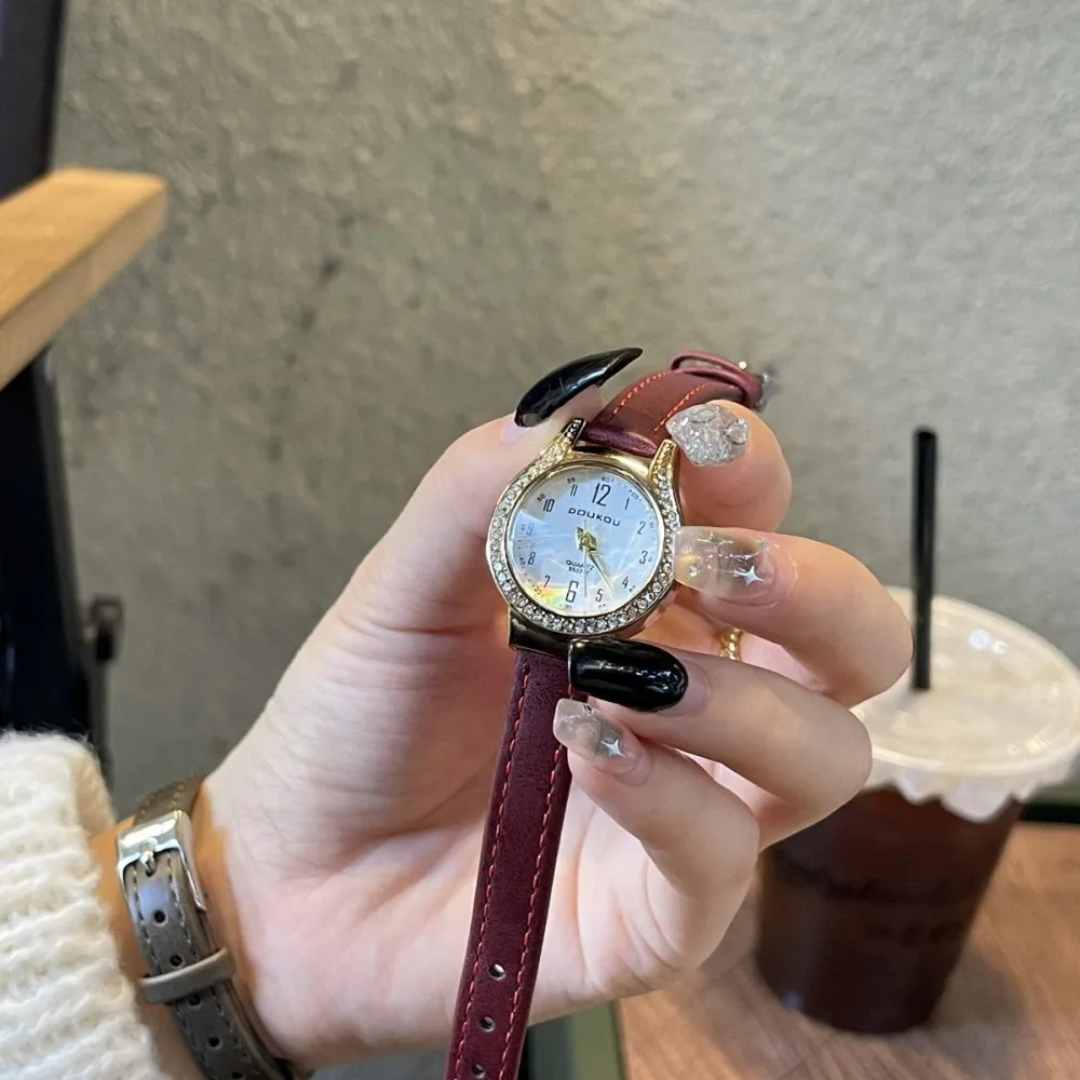 Hand holding a watch with a red strap in front of a blurred background