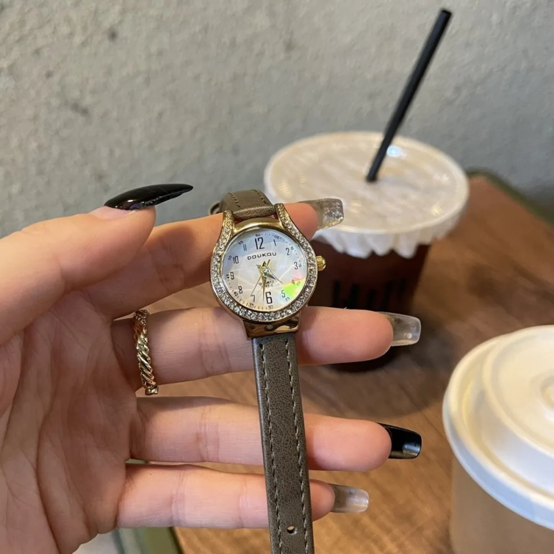 Hand holding a decorative watch with a gray strap in a blurred background of a table with drinks.