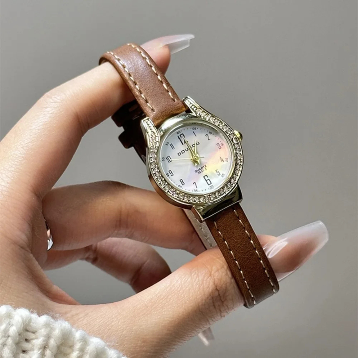 Hand holding a watch with a brown leather strap and decorative case against a neutral background