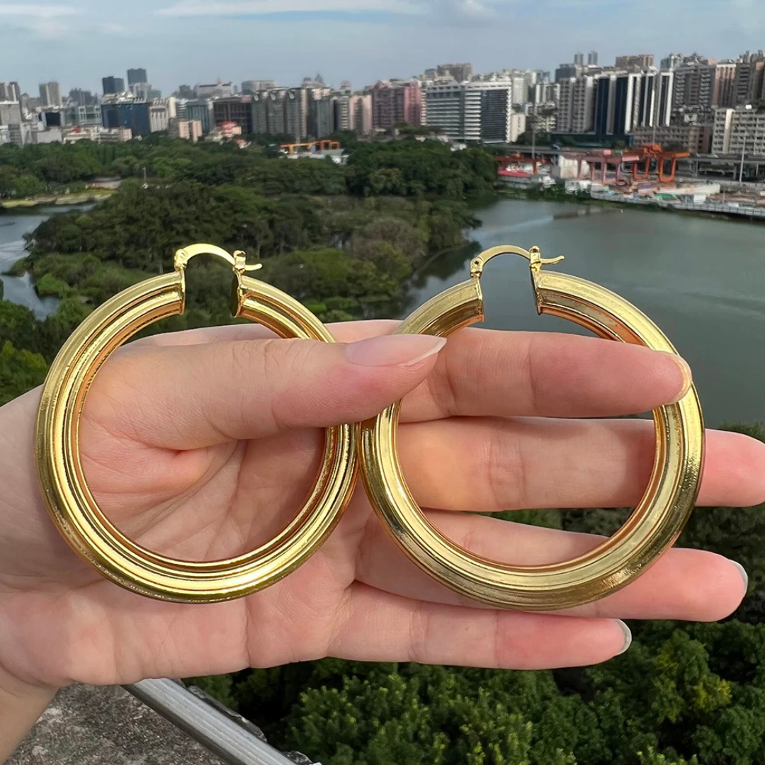 Gold hoop earrings held in front of a cityscape with trees and water.