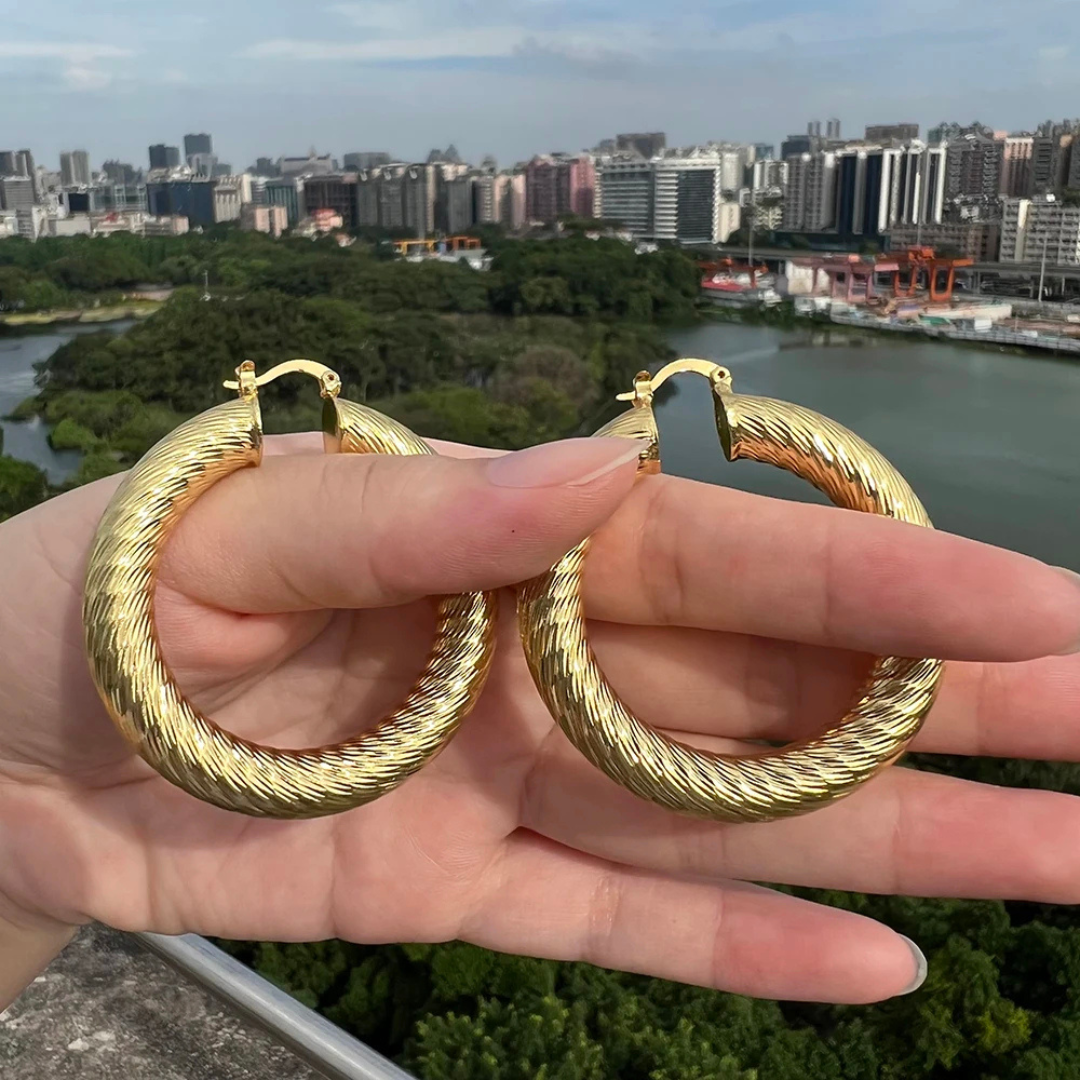 Gold hoop earrings held in front of a cityscape with buildings and water.