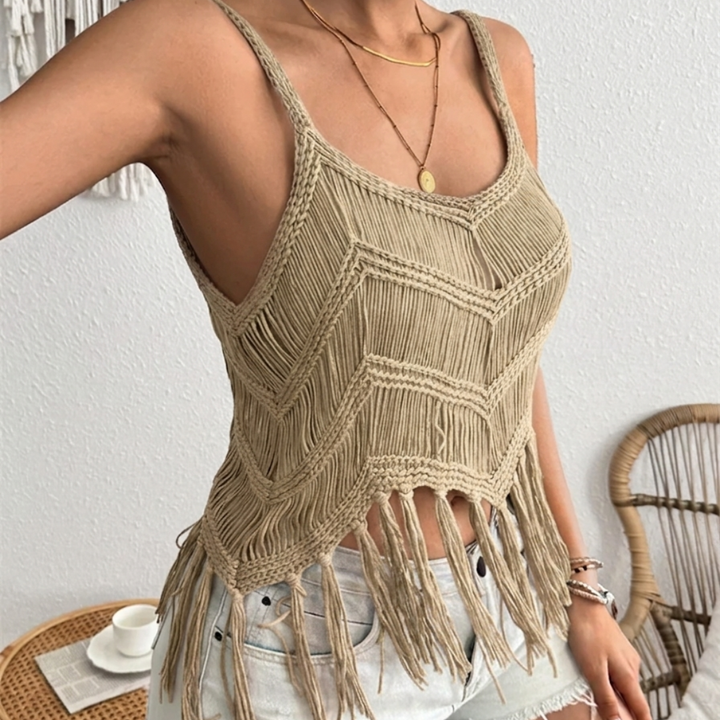 Beige macrame top with fringe details worn by a person, with a white wall and wicker chair in the background.