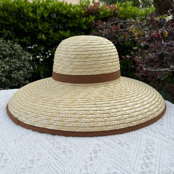 Beige straw hat with brown band on a white surface with greenery in the background
