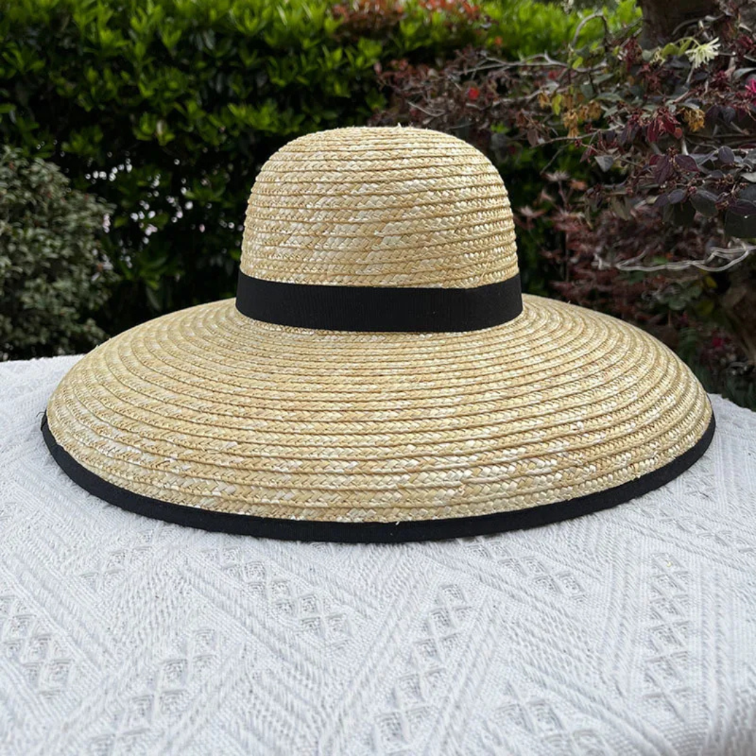 Beige straw hat with black band on a white surface with greenery in the background