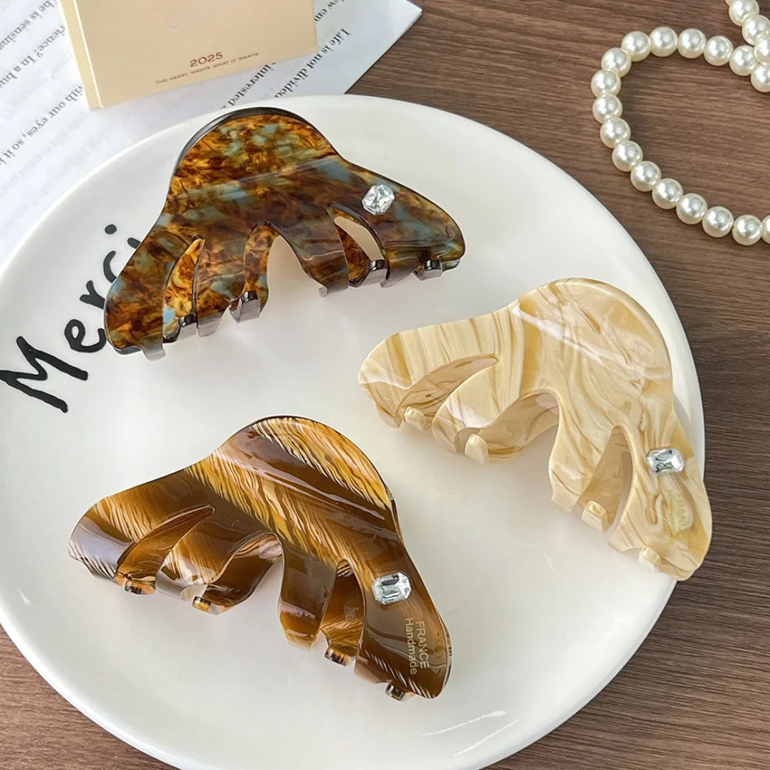 Three tortoiseshell-style hair clips on a white plate with 'Merci' text, pearl necklace in the background.