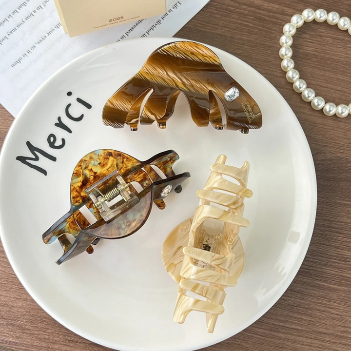 Three tortoiseshell hair clips on a white plate with 'Merci' written on it, next to a pearl necklace.